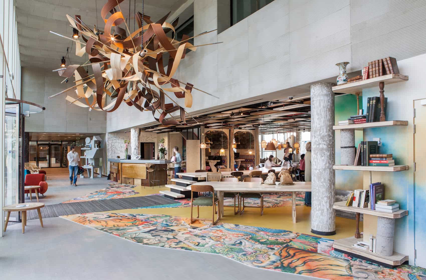 Moderne kantoorhal met een kleurrijk tapijt met patronen, een grote houten kroonluchter, boekenrekken en mensen die op de achtergrond werken.