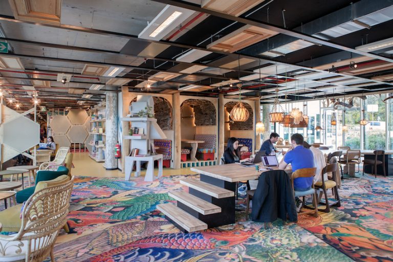 Moderne coworkingruimte met een kleurrijk tapijt met patronen, houten tafels, zitnissen met hanglampen en mensen die op laptops werken.